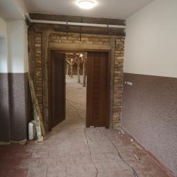 P.W.KOWAL Daniel Kowalski - Wnętrze w trakcie remontu: widoczne niedokończone ściany z cegły, nowa drewniana rama drzwiowa z częściowo zamontowanymi drzwiami, odsłonięta konstrukcja dachu w tle i porozrzucane kable na podłodze.