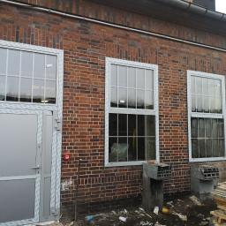 P.W.KOWAL Daniel Kowalski - Murowana ściana budynku z trzema oknami z białymi ramami i szarymi drzwiami wejściowymi. Widoczne ślady po montażu, w tym pozostałości pianki montażowej.