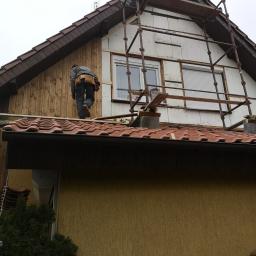P.W.KOWAL Daniel Kowalski - Pracownik na dachu domu w trakcie ocieplania elewacji, widoczne rusztowanie, płyty izolacyjne oraz deski elewacyjne w trakcie montażu.
