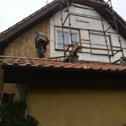 Pracownik na dachu domu w trakcie ocieplania elewacji, widoczne rusztowanie, płyty izolacyjne oraz deski elewacyjne w trakcie montażu.