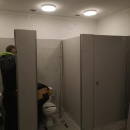 P.W.KOWAL Daniel Kowalski - Instalacja kabin sanitarnych w toalecie publicznej: widoczne kabiny z szarego tworzywa, narzędzia pracy i instalator w trakcie montażu.