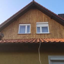 P.W.KOWAL Daniel Kowalski - Pracownik siedzi na dachu z dachówkami i deskami elewacyjnymi, przygotowując się do dalszych prac, widoczne okna z roletami i komin