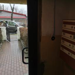 P.W.KOWAL Daniel Kowalski - Widok z klatki schodowej przez otwarte drzwi na zewnątrz, gdzie widać chodnik z czerwonej kostki, furtkę, zaparkowany samochód i budynek z czerwonym dachem.