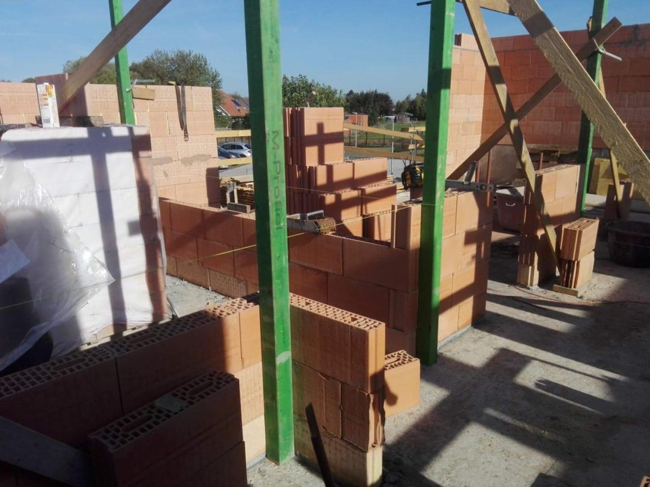 Wznoszenie ścian zewnętrznych z czerwonej cegły ceramicznej, widoczne zielone stemple budowlane i drewniane podpory stabilizujące konstrukcję, etap budowy domu jednorodzinnego.