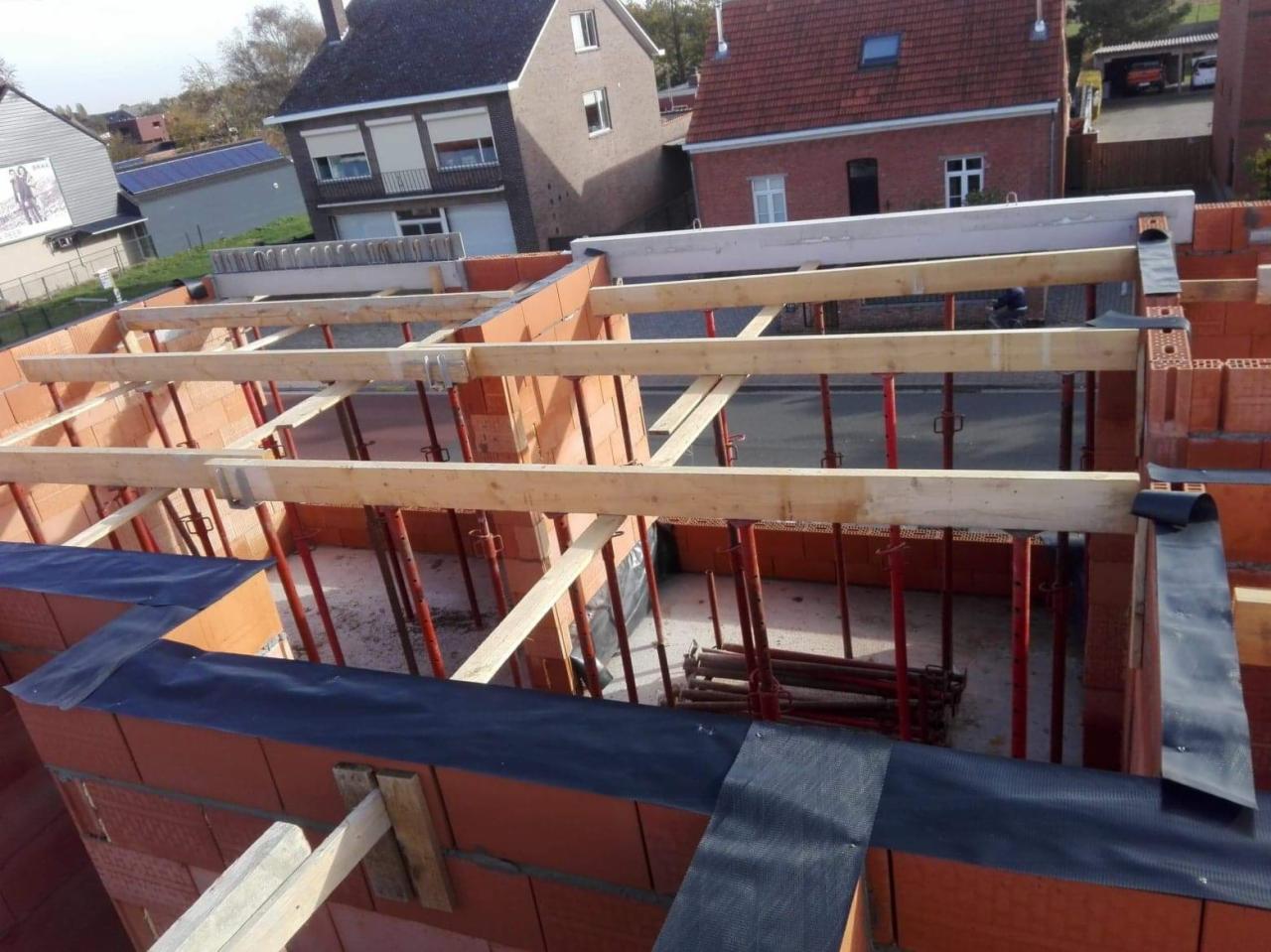 Wznoszenie murów z czerwonej cegły, widoczne stalowe podpory i drewniane belki szalunkowe, czarna folia ochronna na krawędziach, w tle domy jednorodzinne.