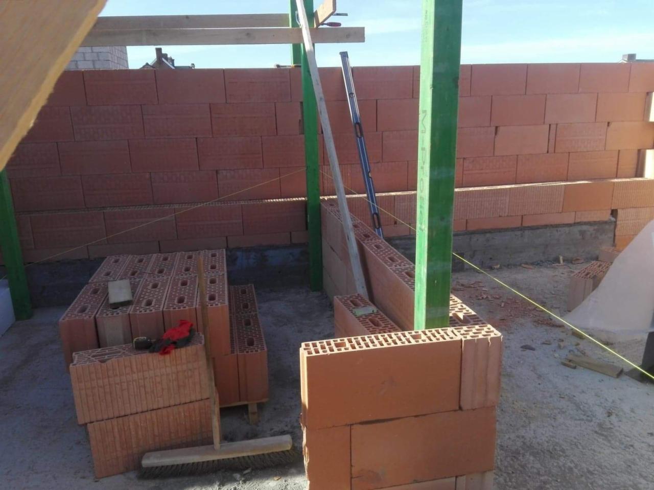 Wznoszenie ściany z czerwonej cegły ceramicznej Porotherm, widoczne zielone stemple podtrzymujące konstrukcję, poziomica i sznurek murarski wyznaczający linię, cegły ułożone na palecie, etap budowy...