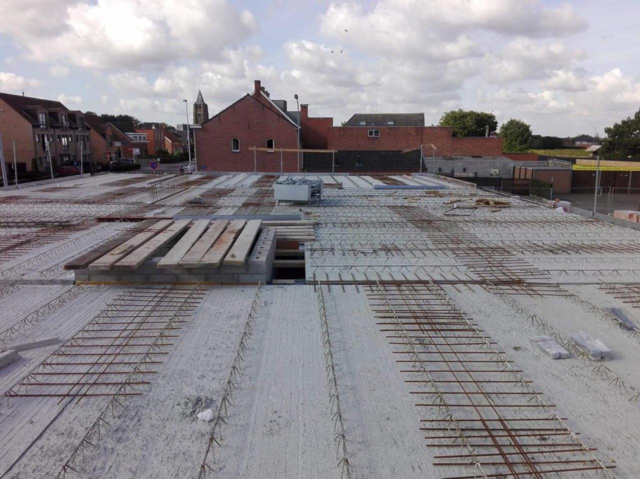 Zbrojenie stropu w trakcie budowy domu jednorodzinnego, widoczne drewniane deski szalunkowe i stalowe pręty zbrojeniowe, w tle zabudowa miejska i pochmurne niebo.