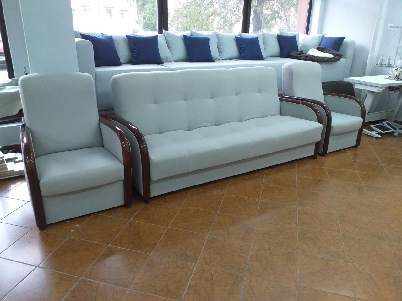 Komplet wypoczynkowy: sofa i dwa fotele z jasną tapicerką i ciemnym drewnianym wykończeniem podłokietników, na tle okna i jasnej ściany.