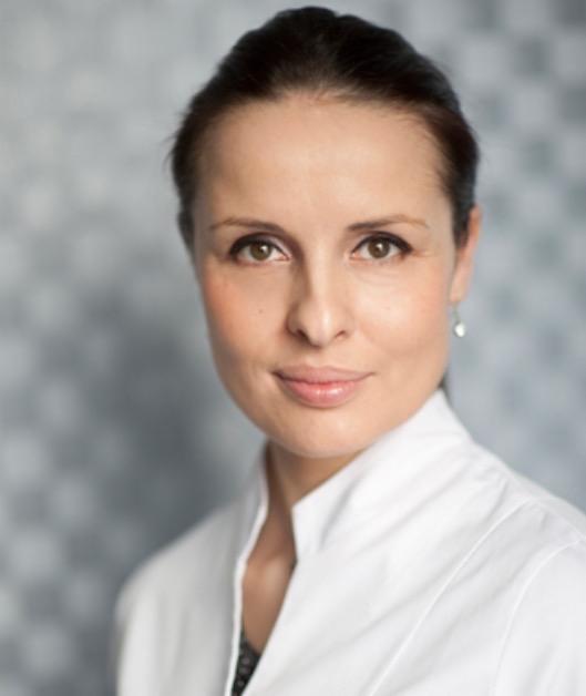 Portret kobiety w białym uniformie medycznym na tle szarej ściany z delikatnym wzorem, z subtelnym kolczykiem w uchu.