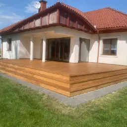 Nowoczesny dom jednorodzinny z tarasem wykonanym z drewna egzotycznego, ułożonym kaskadowo z trzema stopniami, otoczony zielonym trawnikiem i kostką brukową.