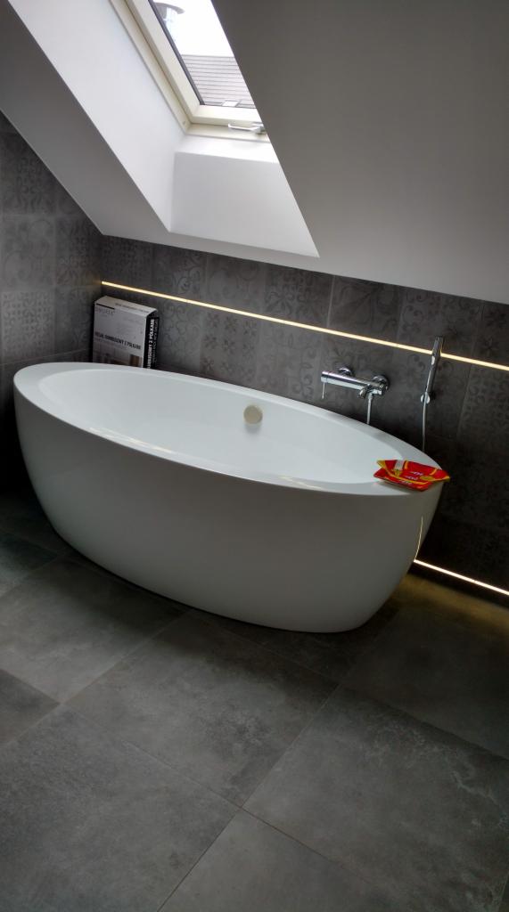 Nowoczesna łazienka na poddaszu z wolnostojącą białą wanną, szarą mozaiką na ścianach, oświetleniem LED i oknem dachowym.