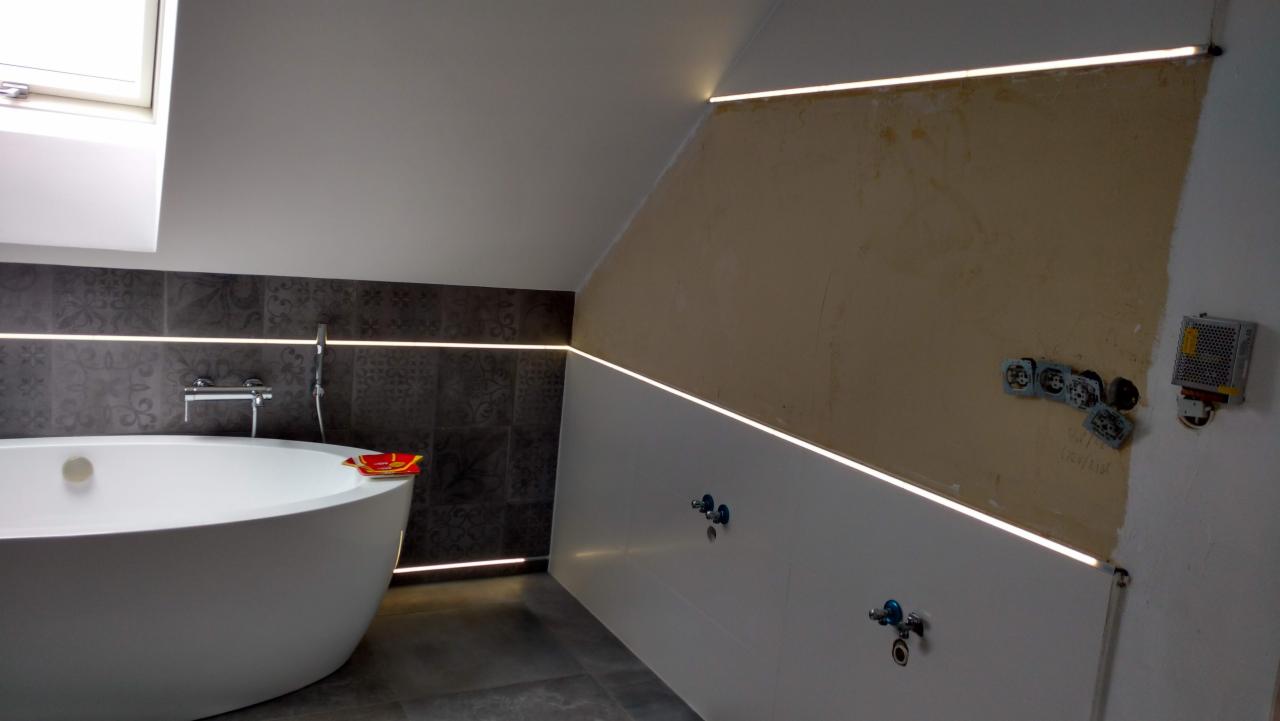 Łazienka w trakcie remontu z podświetleniem LED na ścianach i suficie, widoczna wanna wolnostojąca, ciemne płytki dekoracyjne oraz fragment ściany przygotowanej do malowania z wystającymi gniazdkami.