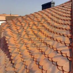 Pomarańczowo-brązowy dach pokryty dachówką bitumiczną w kształcie łuski, widok z góry, w tle komin i zabudowa miejska.