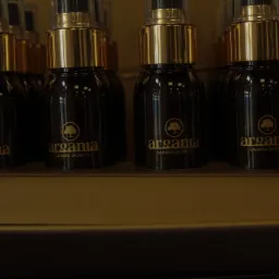 Rząd butelek z ciemnego szkła z atomizerami i złotymi nakrętkami, oznaczonych logo 'argania - SAHARA SECRETS', ustawionych na półce.