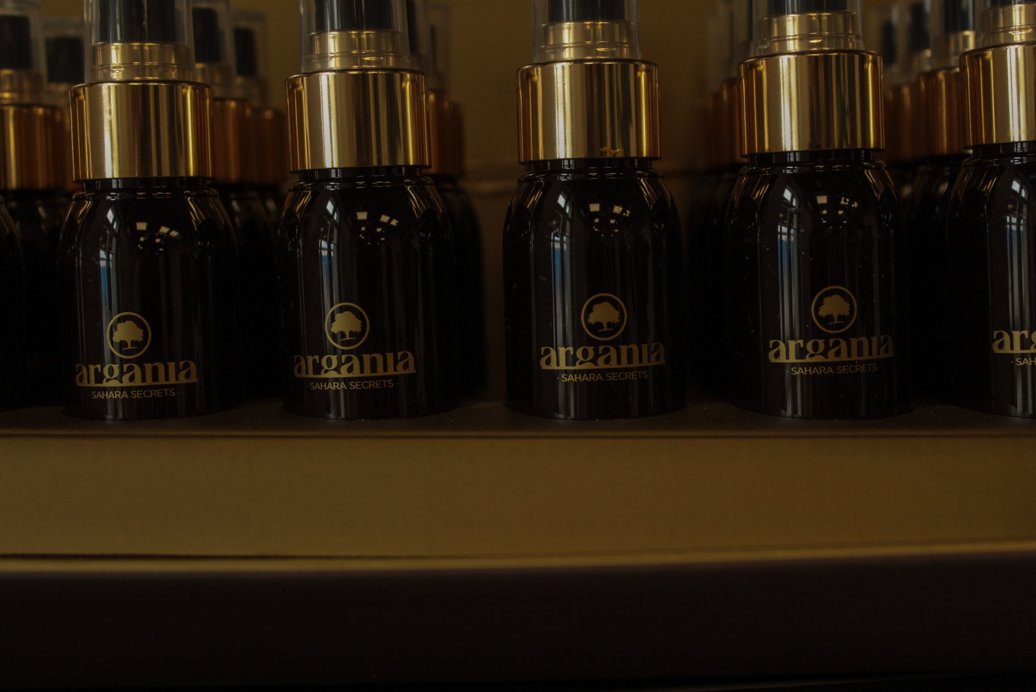 Rząd butelek z ciemnego szkła z atomizerami i złotymi nakrętkami, oznaczonych logo 'argania - SAHARA SECRETS', ustawionych na półce.