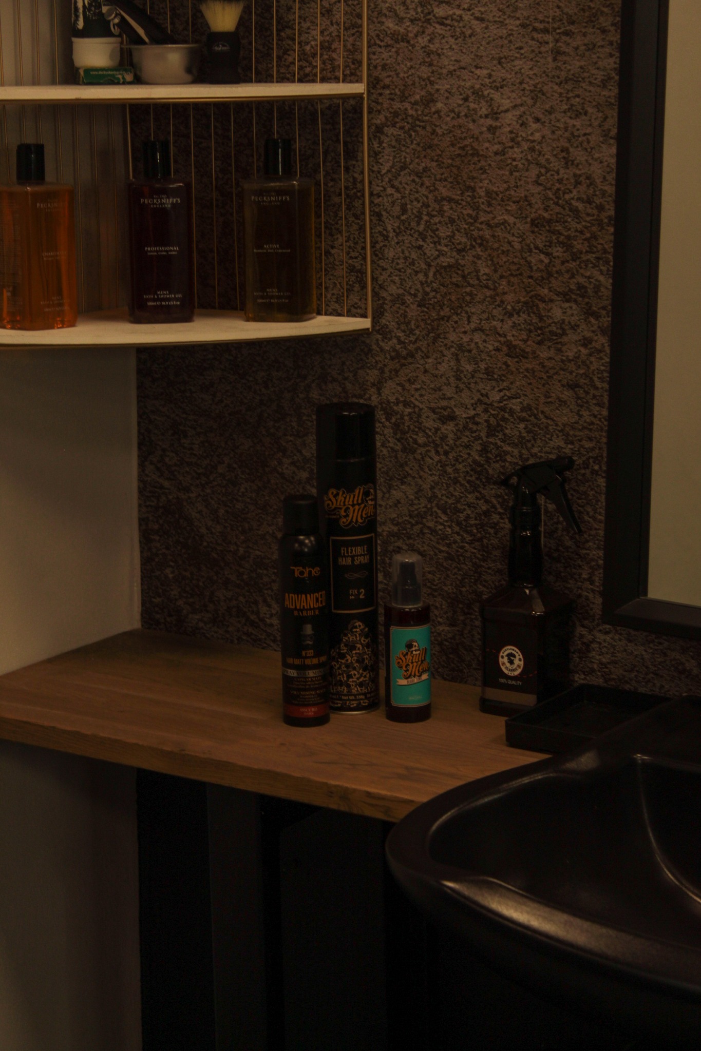 Półka z kosmetykami i akcesoriami fryzjerskimi: płyny do kąpieli Pecksniff's, spray do włosów Skull Men, Tahe Advanced Barber, spryskiwacz fryzjerski i umywalka w salonie fryzjerskim.