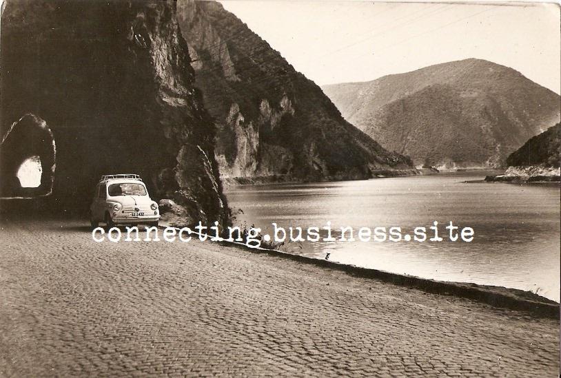 Czarno-białe zdjęcie przedstawia mały, stary samochód osobowy z bagażnikiem na dachu, wyjeżdżający z tunelu skalnego nad brzegiem jeziora, w tle góry.