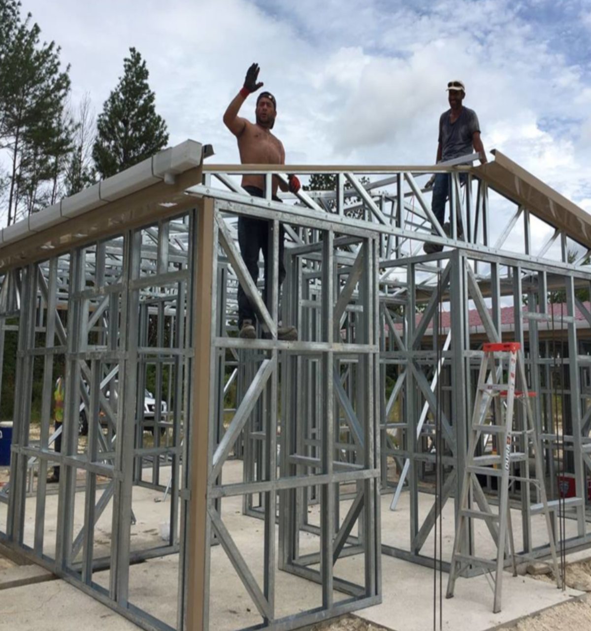 Konstrukcja domu ze stalowym szkieletem w trakcie budowy; jeden z robotników macha ręką z góry, drugi pracuje na dachu; widoczna drabina i betonowa podstawa.
