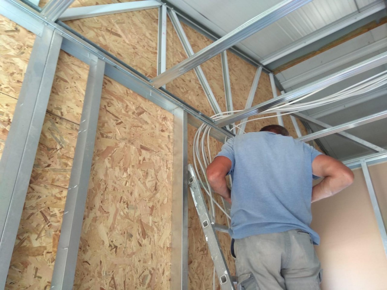 Konstrukcja szkieletowa domu w trakcie budowy: widoczne metalowe profile, płyta OSB, instalacja elektryczna oraz pracownik na drabinie.