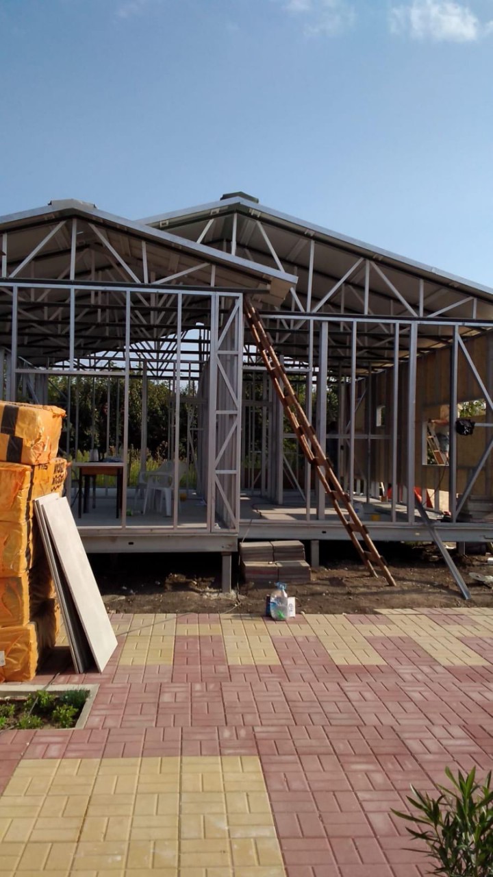 Konstrukcja szkieletowa domu z widocznymi elementami stalowymi, drewnianą drabiną opartą o ścianę, paczkami izolacji termicznej oraz ułożoną kostką brukową na zewnątrz.