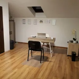 Gabinet z biurkiem, laptopem, krzesłem i dyplomami na ścianie, oświetlony światłem z okna dachowego.