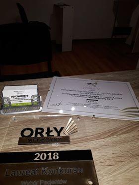 Dyplom, wizytówki w metalowym podajniku i nagroda Orły Medycyny 2018 na drewnianym biurku, w tle krzesło i szafka.