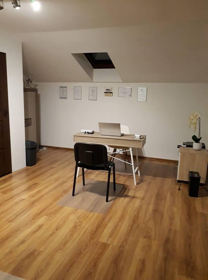 Gabinet z biurkiem, laptopem, krzesłem i dyplomami na ścianie, oświetlony światłem z okna dachowego.