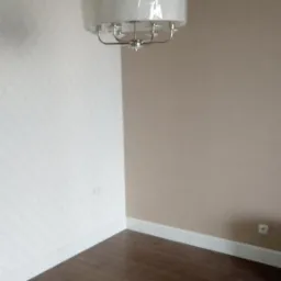 Pomalowane ściany w pokoju, jedna w kolorze beżowym, druga pokryta tapetą z delikatnym wzorem, drewniana podłoga, białe listwy przypodłogowe i sufitowe, oraz nowoczesny, wiszący żyrandol z abażurem.