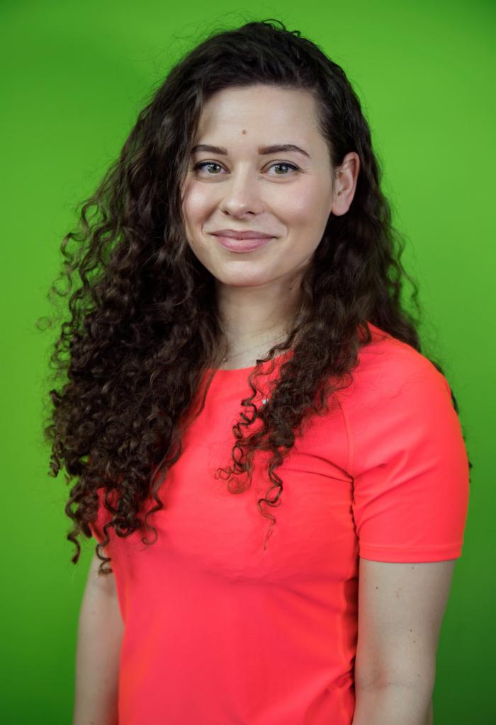 Portret uśmiechniętej kobiety z kręconymi włosami w neonowej pomarańczowej koszulce na jednolitym zielonym tle.