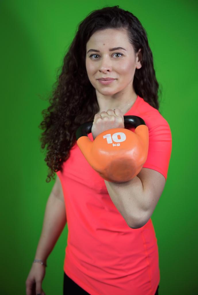 Kobieta z kręconymi włosami, w neonowej koszulce, trzyma pomarańczowy kettlebell 10 kg na tle zielonego ekranu.