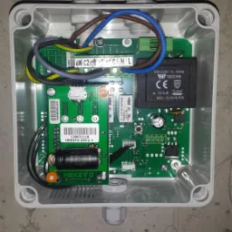 Otwarta szara skrzynka elektryczna z widoczną zieloną płytką drukowaną, przewodami i komponentami elektronicznymi, zamontowana na szarej, surowej ścianie.