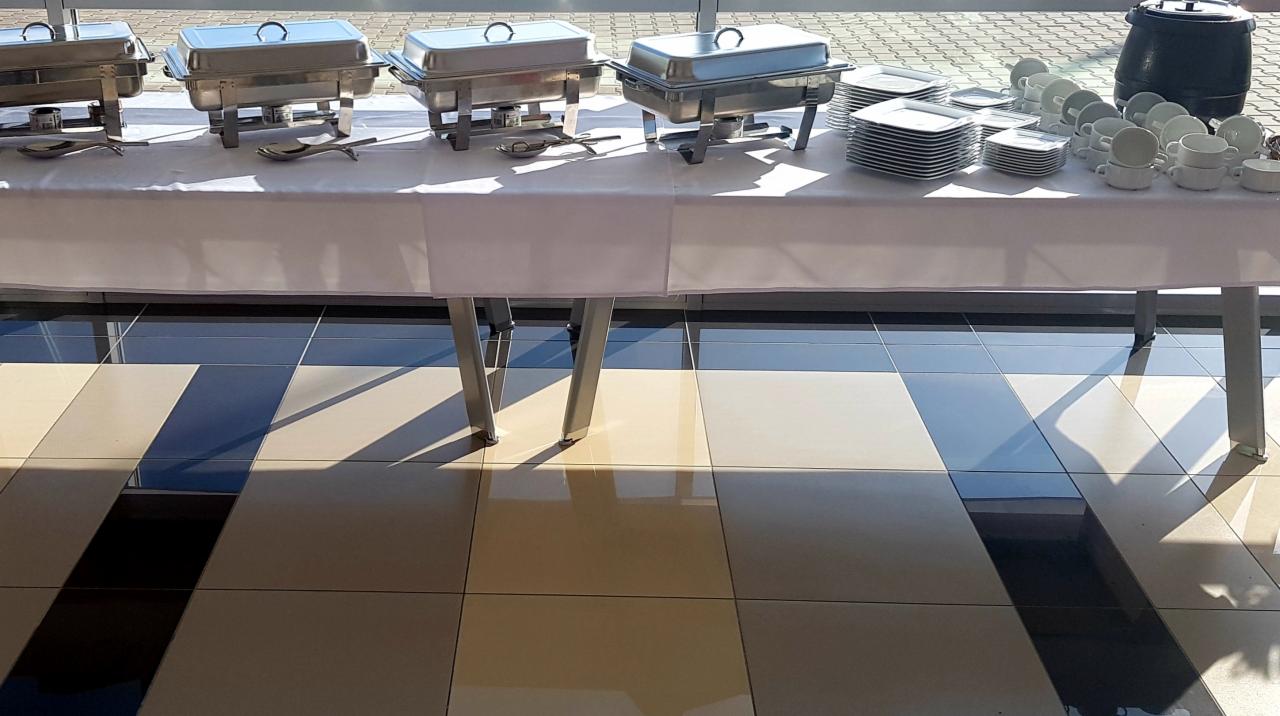 Przygotowany stół cateringowy z podgrzewaczami, stertami białych talerzy i filiżanek, oświetlony naturalnym światłem wpadającym przez okno, na tle wzorzystej podłogi.