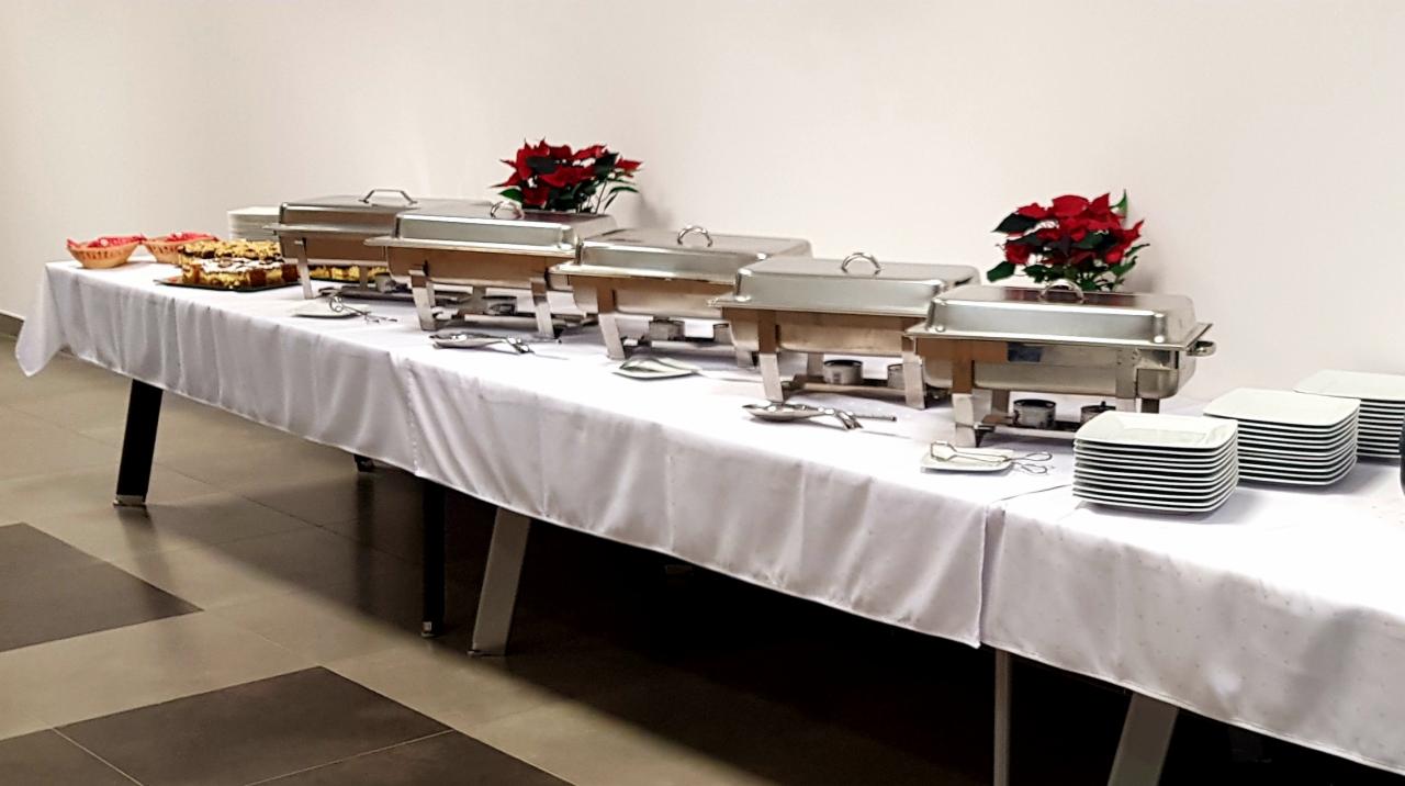 Stół cateringowy przykryty białym obrusem z podgrzewaczami, talerzami i drobnymi przekąskami, udekorowany czerwonymi kwiatami poinsecji.