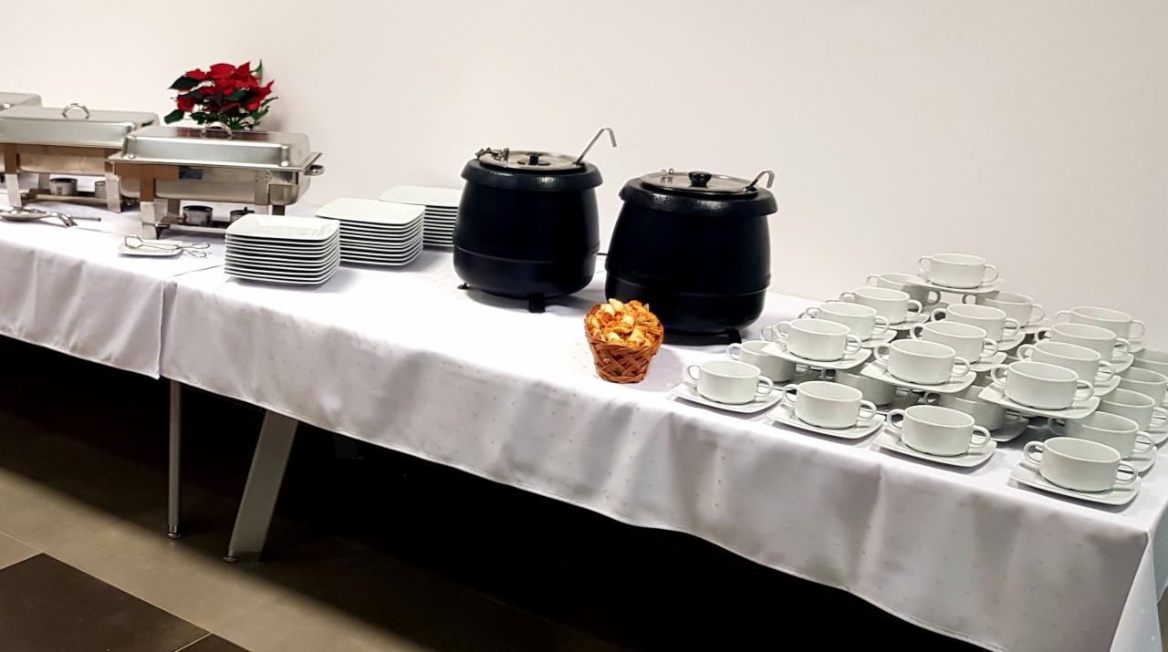 Stół cateringowy z białym obrusem, metalowe bemary z podgrzewaczami, stosy kwadratowych talerzy, dwa czarne kotły z chochlami, koszyk z pieczywem i piramida z białych filiżanek na spodkach.