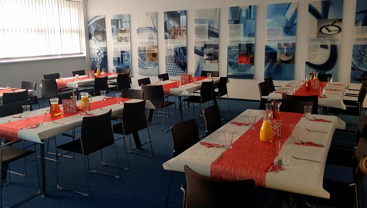 Sala konferencyjna przygotowana do uroczystego obiadu z równo ustawionymi stołami, białymi obrusami z czerwonymi bieżnikami, szklankami i sztućcami, dzbankami z sokiem pomarańczowym oraz czarnymi...