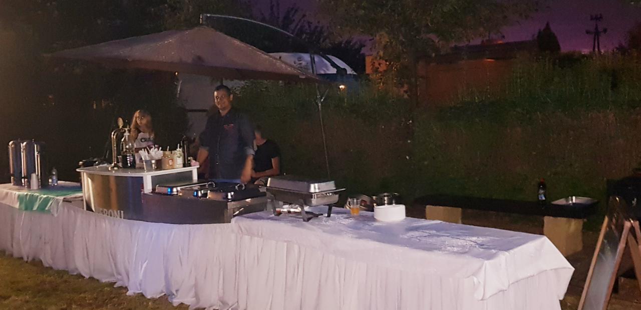 Zewnętrzny bufet z białym obrusem, grillem, podgrzewaczami, dystrybutorem napojów i obsługą, oświetlony w nocy, z widocznym pojazdem w tle.