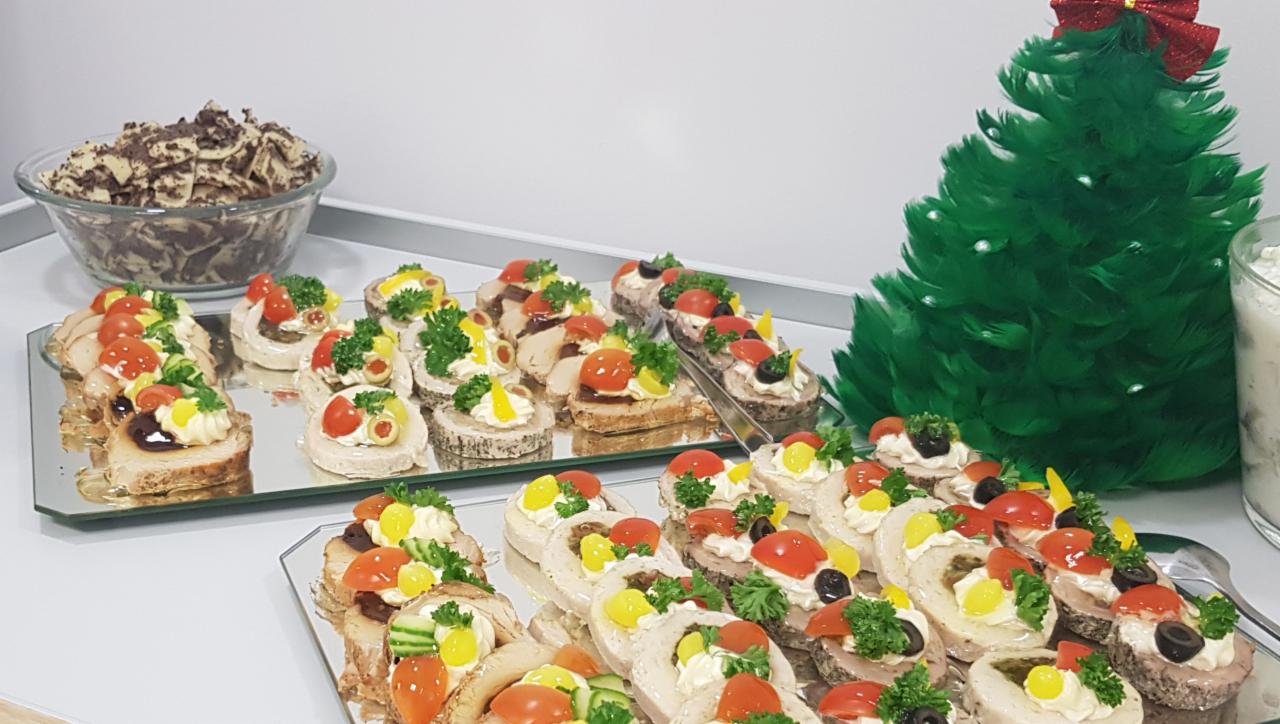 Świąteczny stół cateringowy z różnorodnymi przekąskami na srebrnych tacach, w tle dekoracyjna choinka z piór z czerwoną kokardą i miska z deserem.