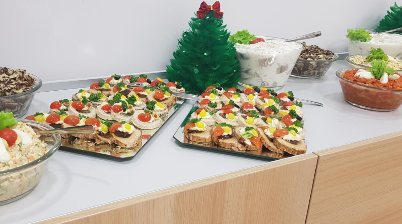 Dekoracyjnie ułożone kanapeczki na srebrnych tacach, sałatki w szklanych misach i mała zielona choinka z czerwonymi bombkami na stole.
