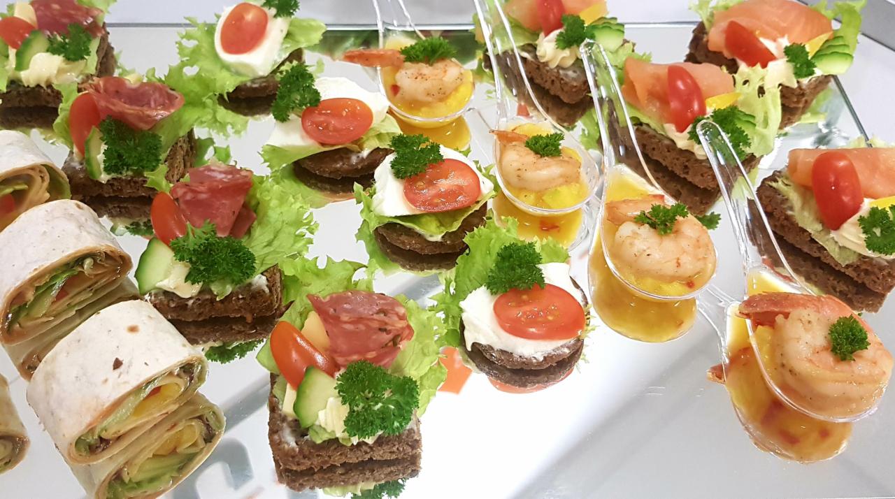 Elegancki zestaw przekąsek na srebrnej tacy: mini tortille z warzywami, kanapeczki na ciemnym pieczywie z wędlinami, serem i warzywami, oraz krewetki w sosie podane w przezroczystych łyżeczkach.