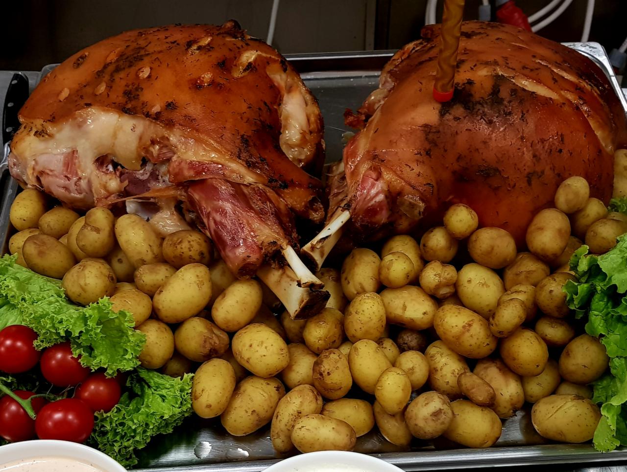 Dwie pieczone golonki na srebrnej tacy, otoczone młodymi ziemniakami, pomidorkami cherry i sałatą, widoczne sosy w miseczkach w dolnej części kadru.