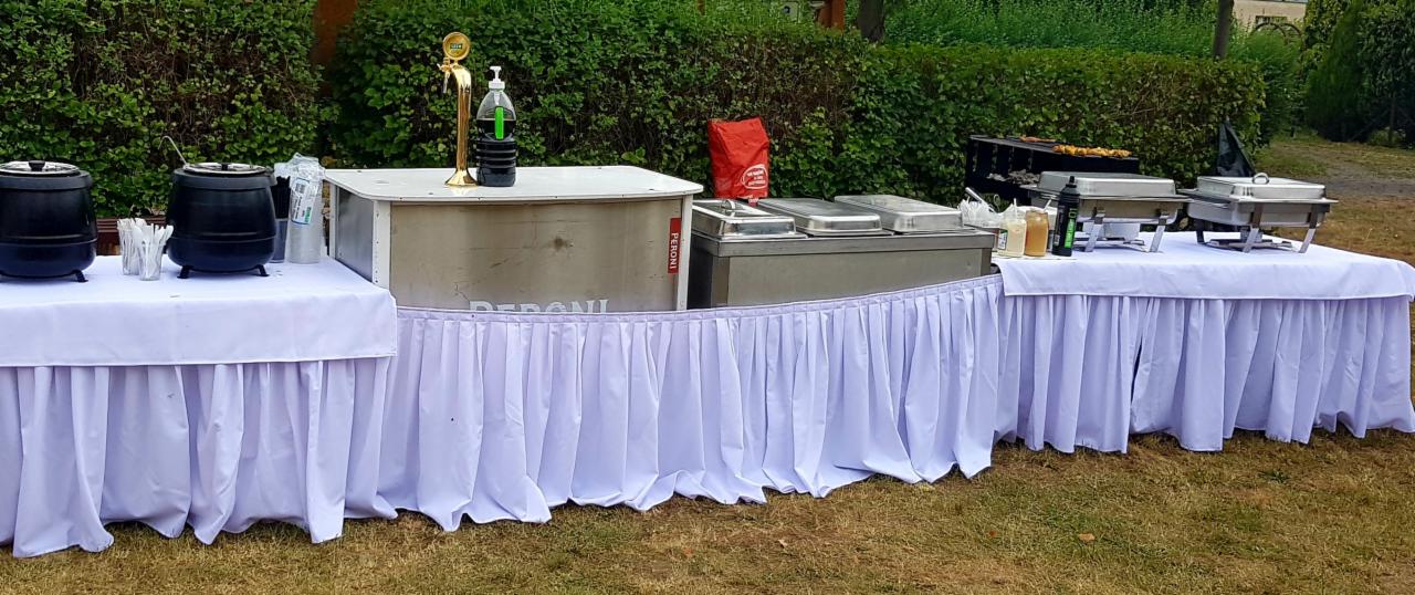 Rozstawione stoły cateringowe z białymi obrusami, na nich podgrzewacze, zupy w kotłach i dystrybutor napojów na tle zieleni.