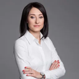 Portret kobiety w białej koszuli z założonymi rękami, na szarym tle, z delikatnym uśmiechem i brązowym paskiem zegarka.