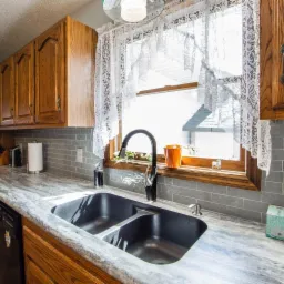 Wykończona kuchnia z drewnianymi szafkami, szarymi płytkami na ścianie i czarnym, dwukomorowym zlewem pod oknem z koronkową firanką.