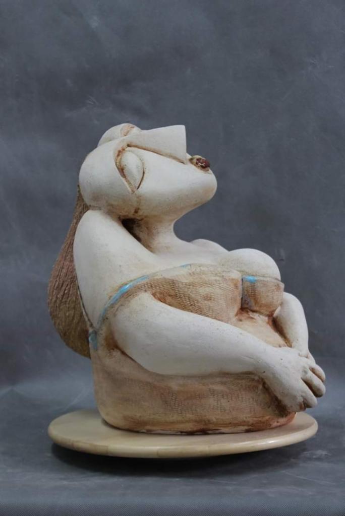 Ręcznie wykonana, ceramiczna rzeźba kobiety siedzącej na okrągłej podstawie, z detalami w kolorze niebieskim, na szarym tle.