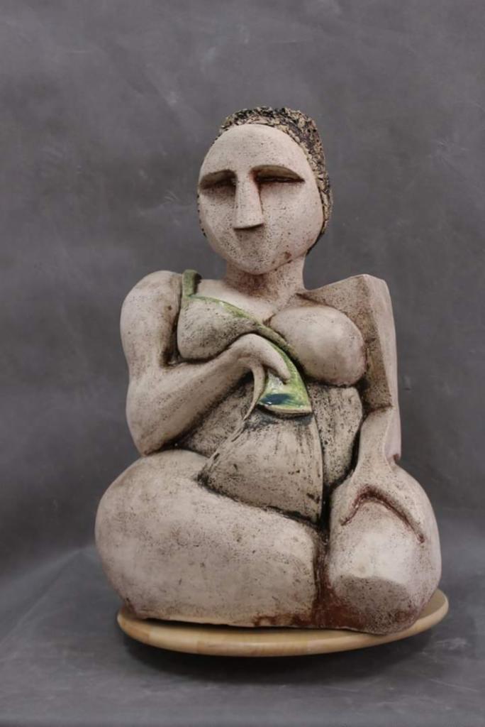 Ceramiczna rzeźba siedzącej kobiety o uproszczonych rysach, z zielonym akcentem na piersiach, na drewnianej podstawie, na tle szarego tła.