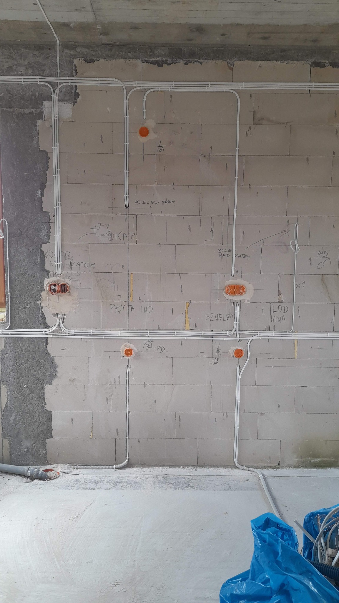 Surowa ściana z betonu komórkowego z widocznymi białymi rurami instalacji elektrycznej prowadzonymi natynkowo, pomarańczowe puszki pod gniazdka i włączniki, oraz odręczne notatki na ścianie.