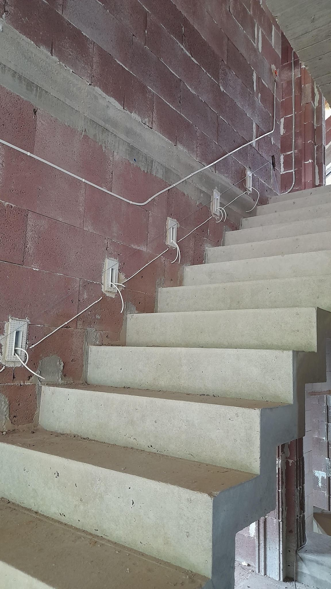Betonowe schody w surowym stanie z widocznymi przewodami elektrycznymi w peszlach, zamontowanymi na ścianie z czerwonej cegły dziurawki. W ścianie przygotowane puszki pod gniazdka elektryczne.