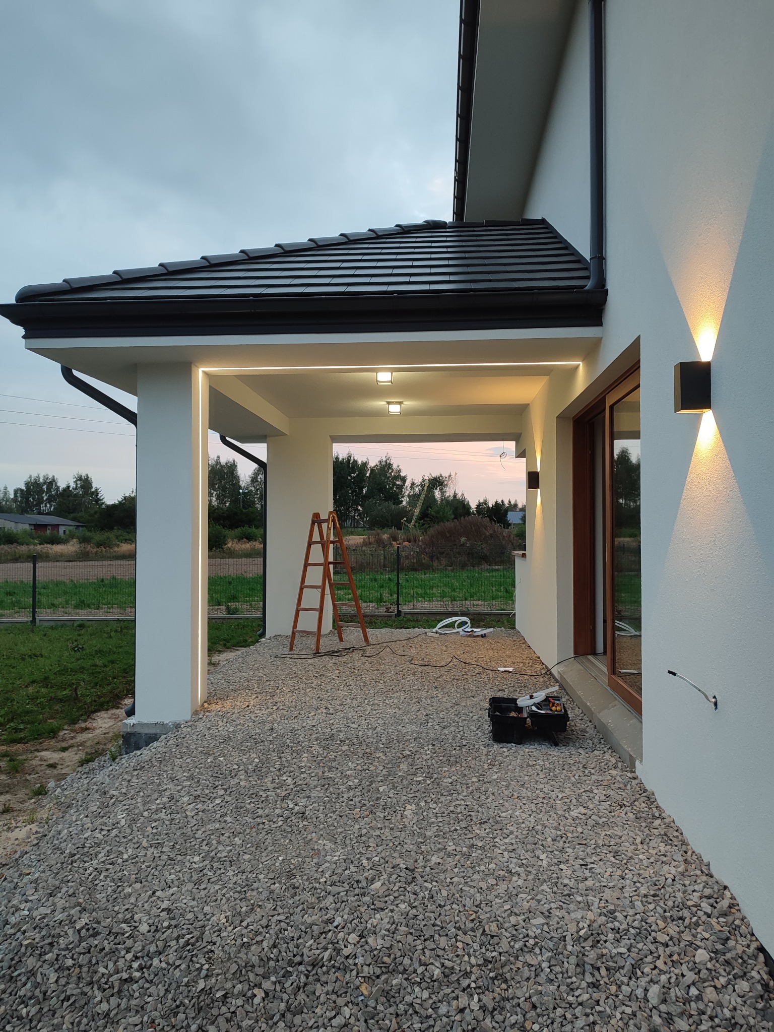 Nowoczesny dom z podświetlanym gankiem i zewnętrznymi lampami ściennymi, drabina i skrzynka z narzędziami na żwirowym podłożu sugerują trwające prace.