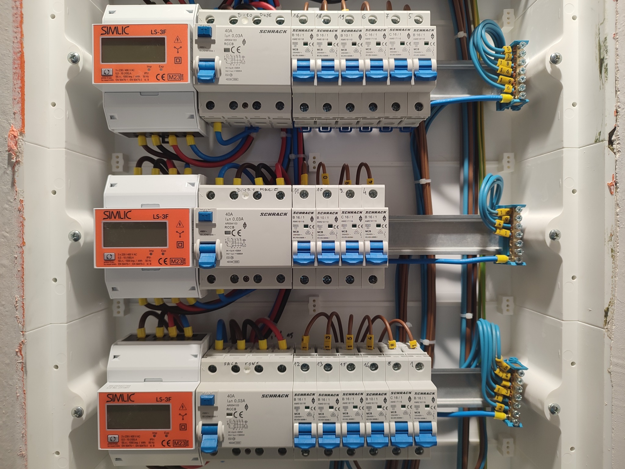 Rozdzielnica elektryczna z zamontowanymi bezpiecznikami Schrack i licznikami Simlic LS-3F, widoczne kable z oznaczeniami i złączki, instalacja po montażu.