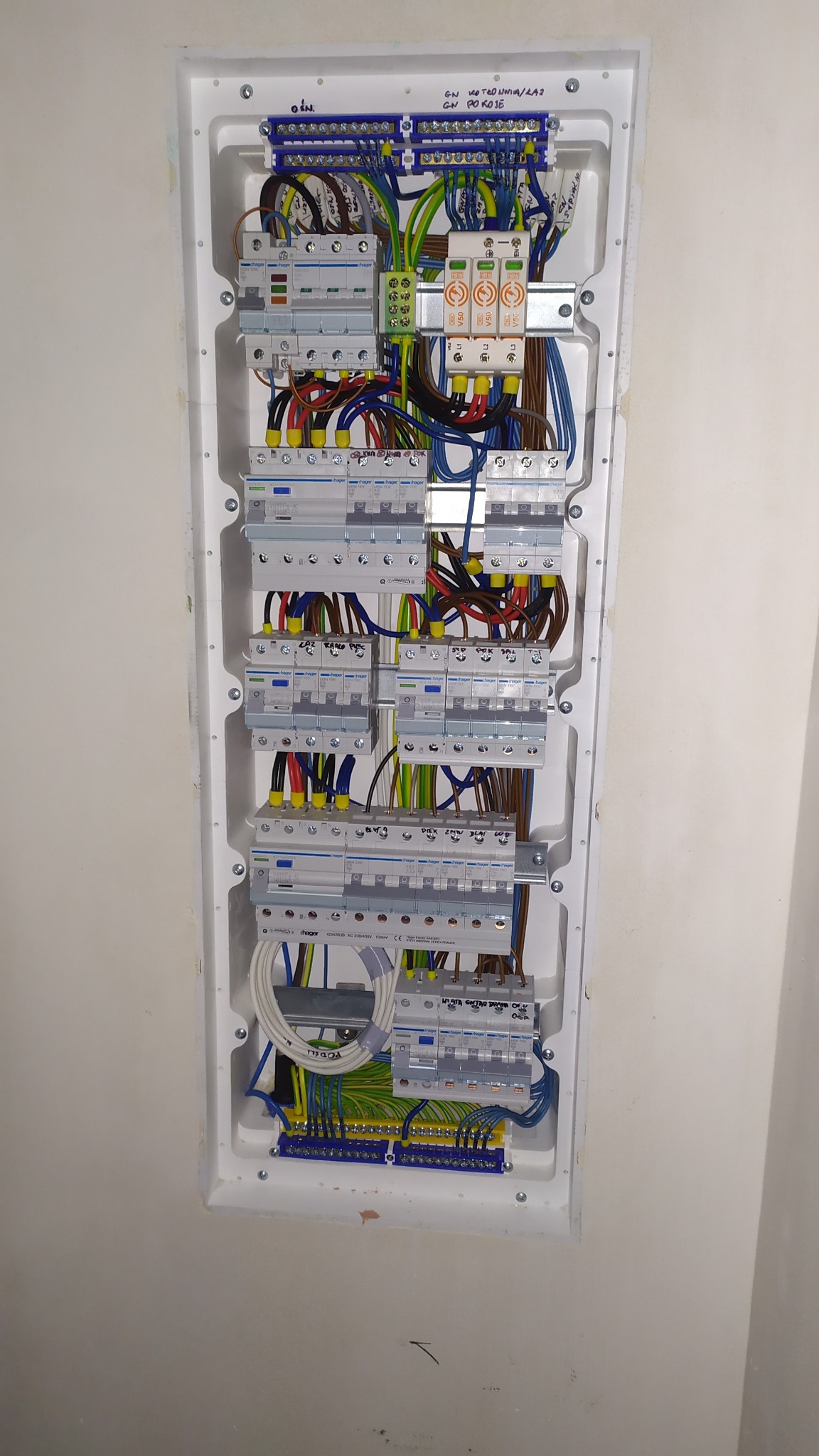 Rozdzielnica elektryczna Hager zamontowana w białej obudowie w ścianie, z widocznymi bezpiecznikami, przewodami o różnych kolorach i oznaczeniach, oraz listwami zaciskowymi.